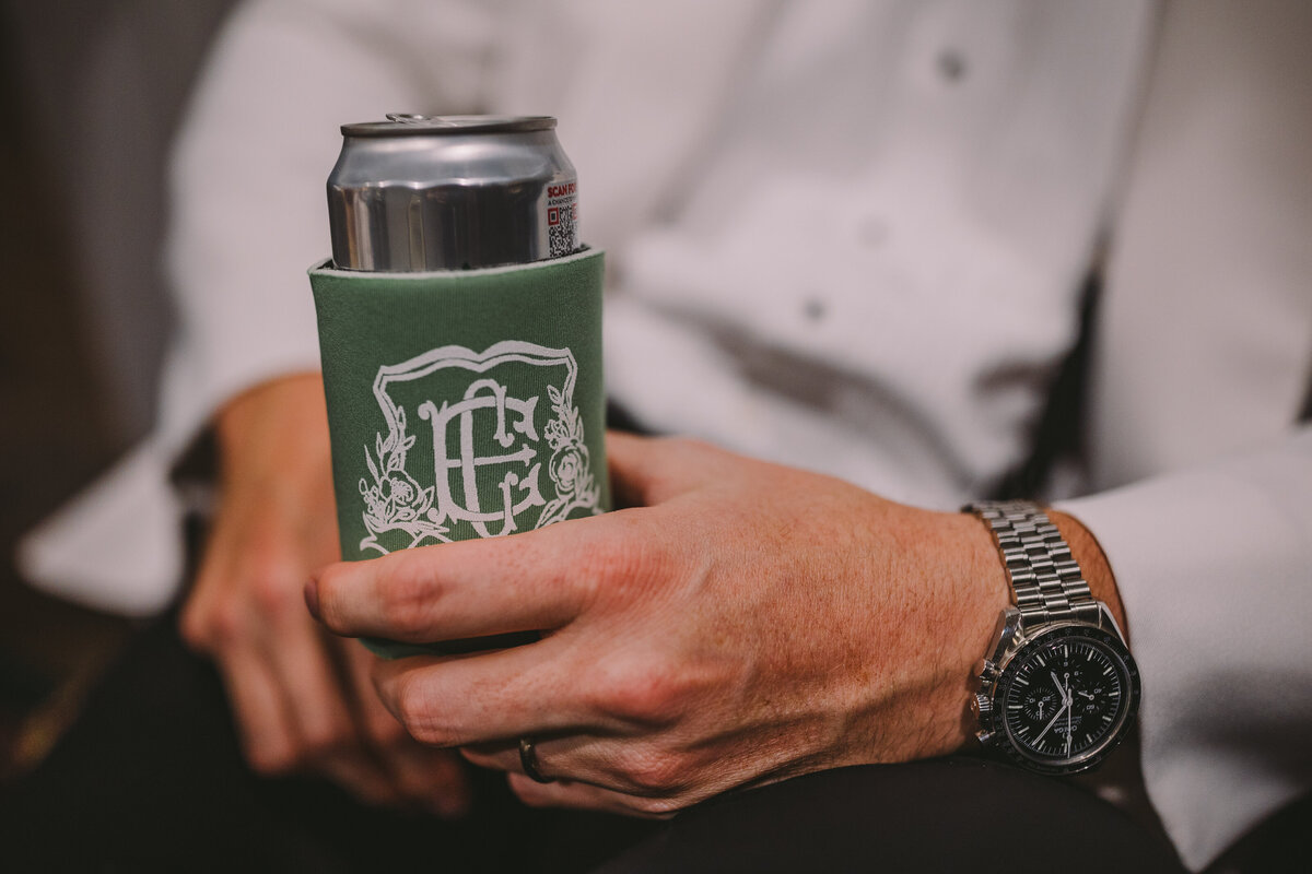 Groom holding a can holder featuring a wedding monogram with florals and the couple's initials.