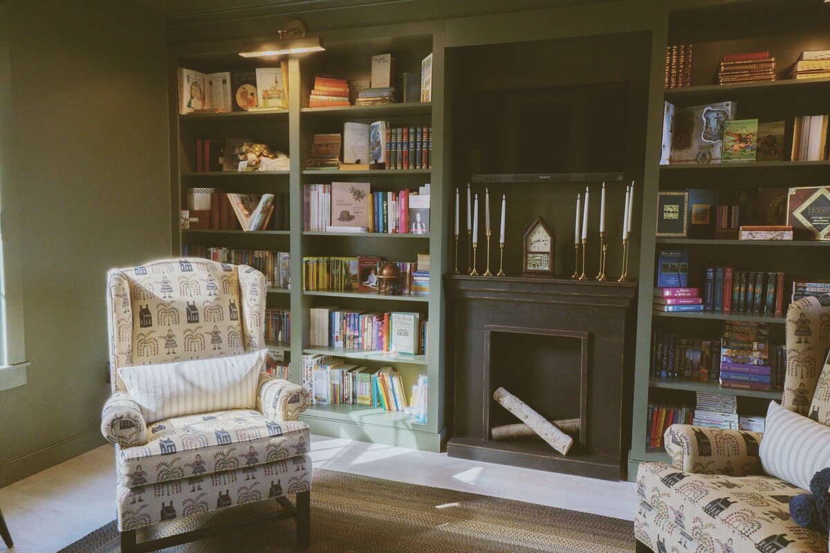 Library with built-ins and faux fireplace with birch logs and two early American textile covered armchairs