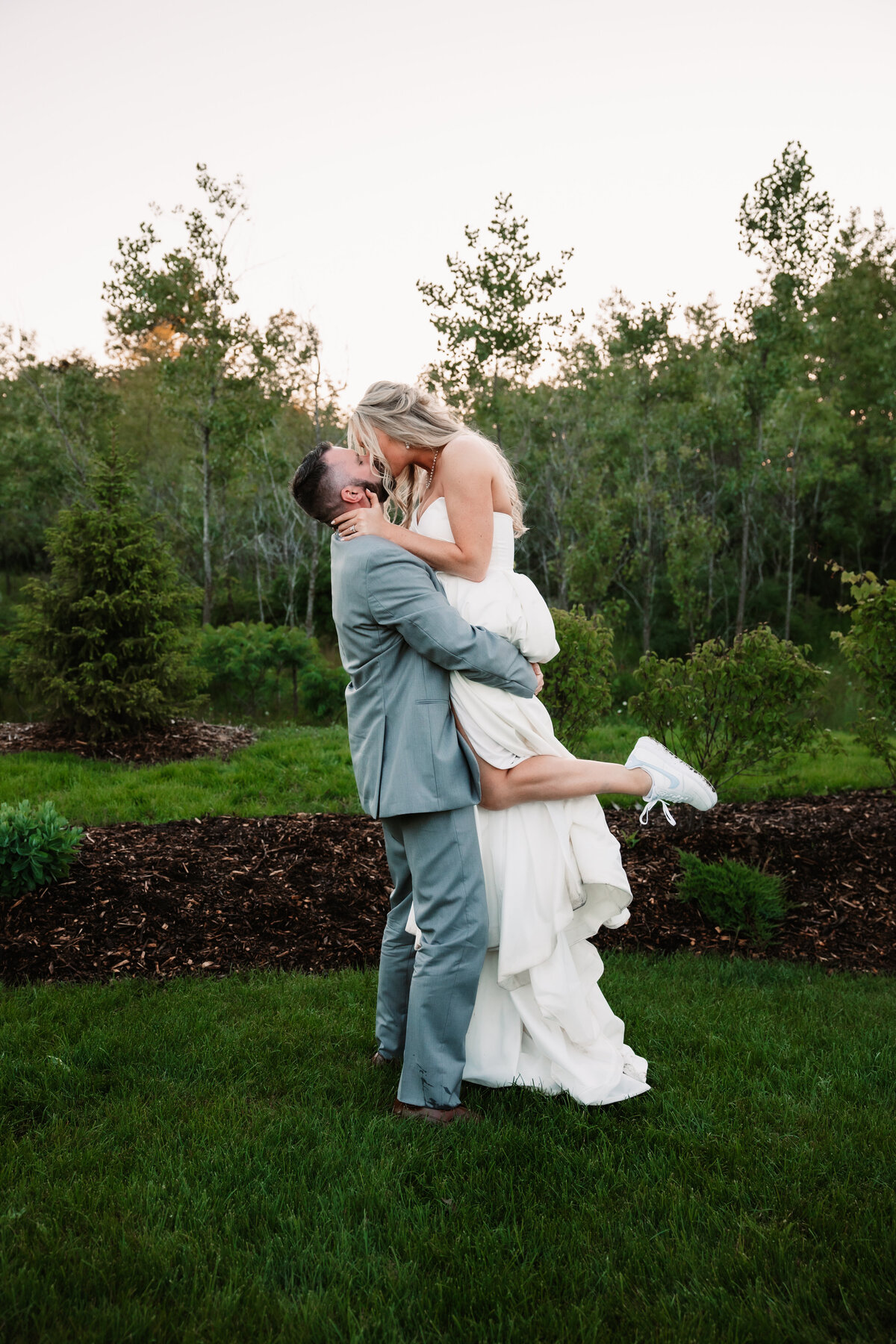 Newlyweds Kiss while he lifts his bride in a garden at sunset