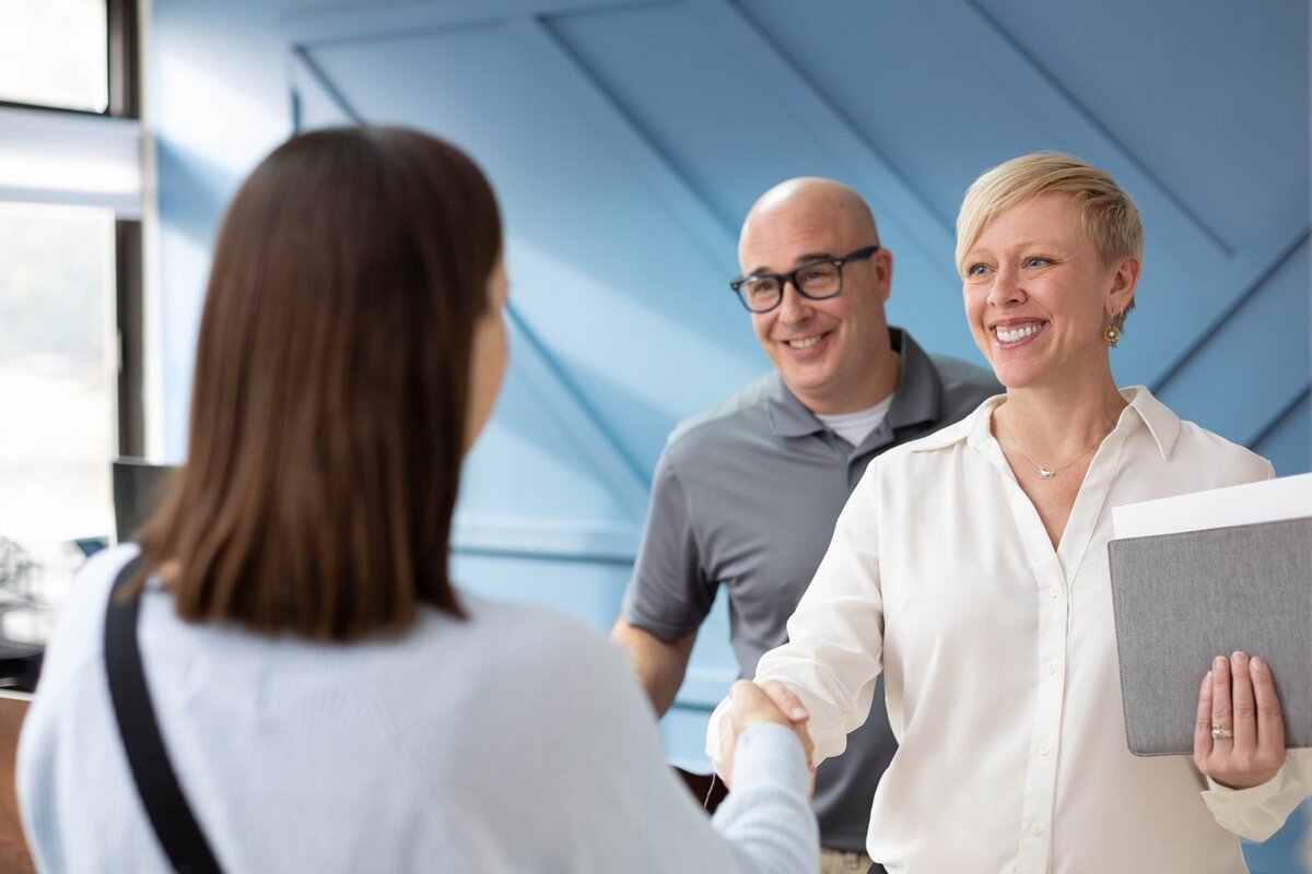 woman with a short haircut smiling and shaking hands with a customer, with a man in glasses standing behind her.