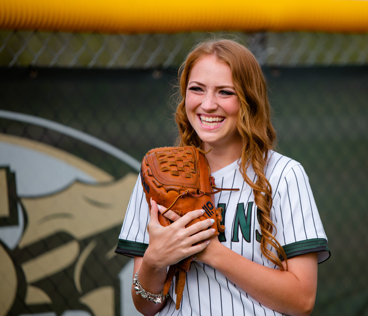 A high school senior stands in a softball outfield near the fence and holds her glove near her chest while smiling.