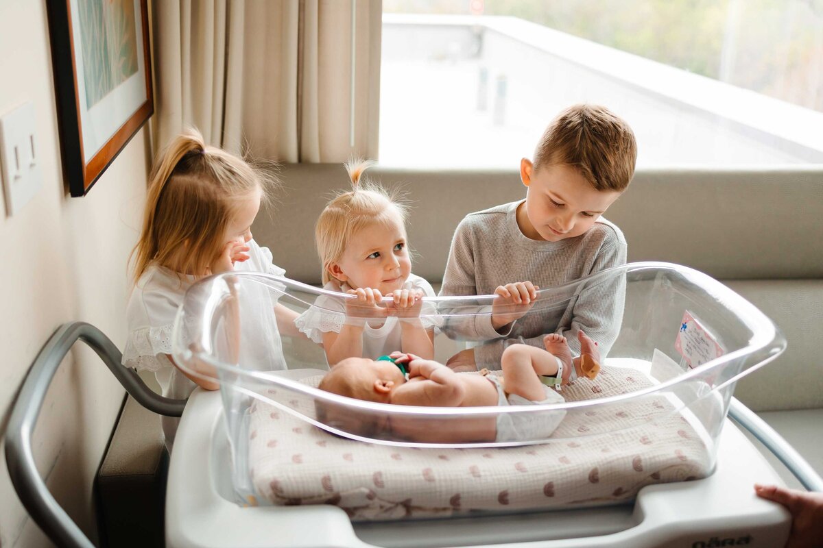 A serene image of a newborn baby peacefully sleeping in a transparent hospital cradle. The baby is surrounded by three older siblings who are gazing at him with love and kindness.