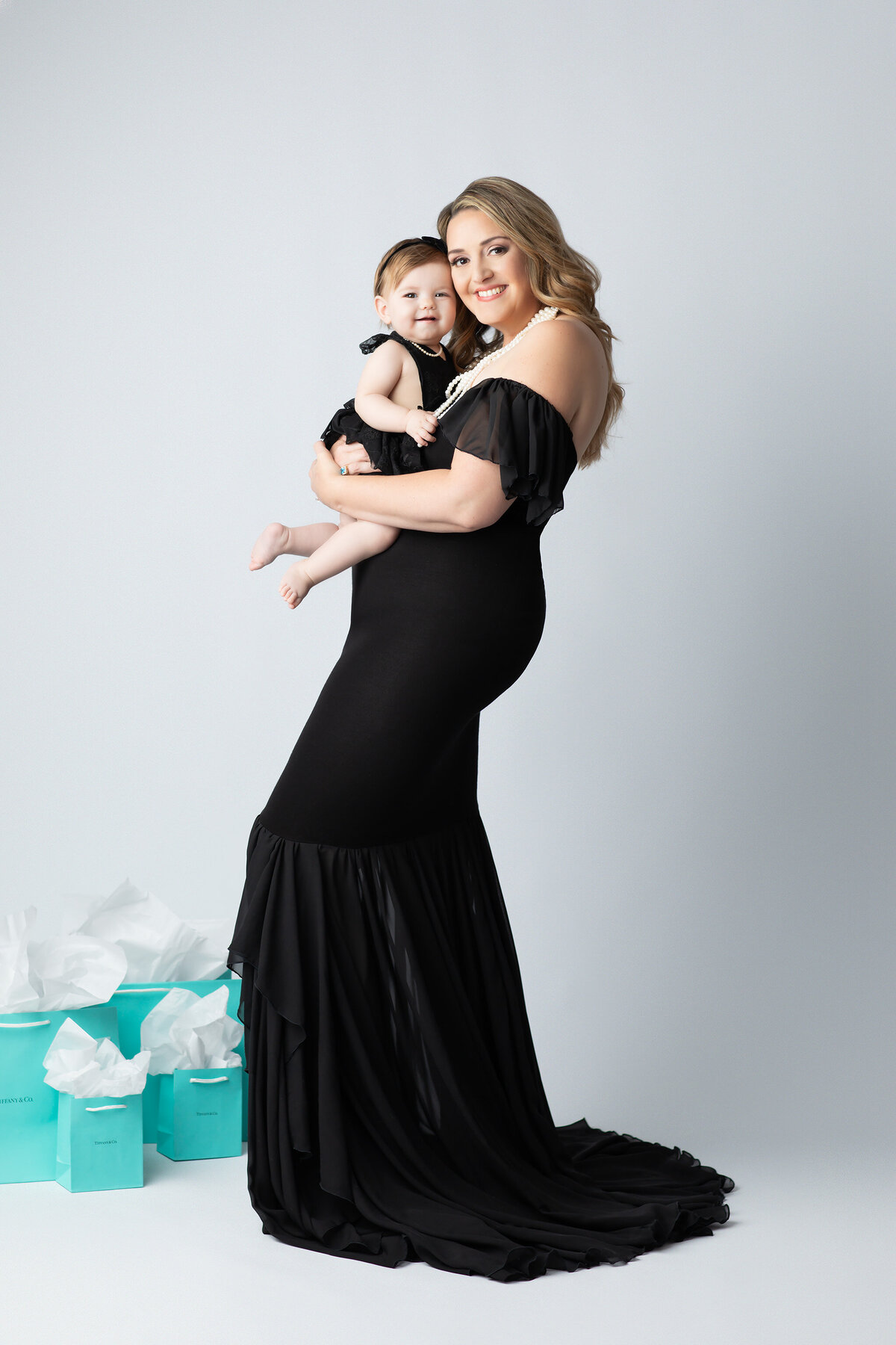 Dallas milestone photographer captures a timeless mother and daughter portrait with both dressed in matching black gowns and pearls, standing beside Tiffany blue gift bags for a chic, fashion-inspired look.