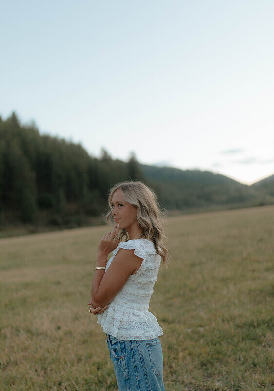Gently touching her face with one hand, a girl looks off to the side for a senior photo. She's standing in a grass field in Idaho Falls, ID.