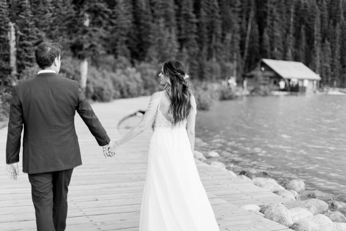 black-and-white-photograph-of-bride-and-groom-walking-at-Lake-Louise-toward-the-old-boat-house