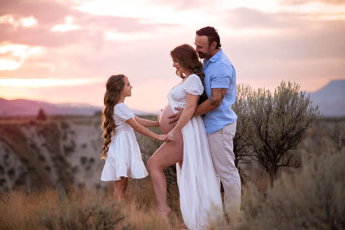 A family of three pose for a Maternity image. There is a soft sunet behind them and the mother and daughter look at eachother while holding hands. Image by brittany Danielle Photography