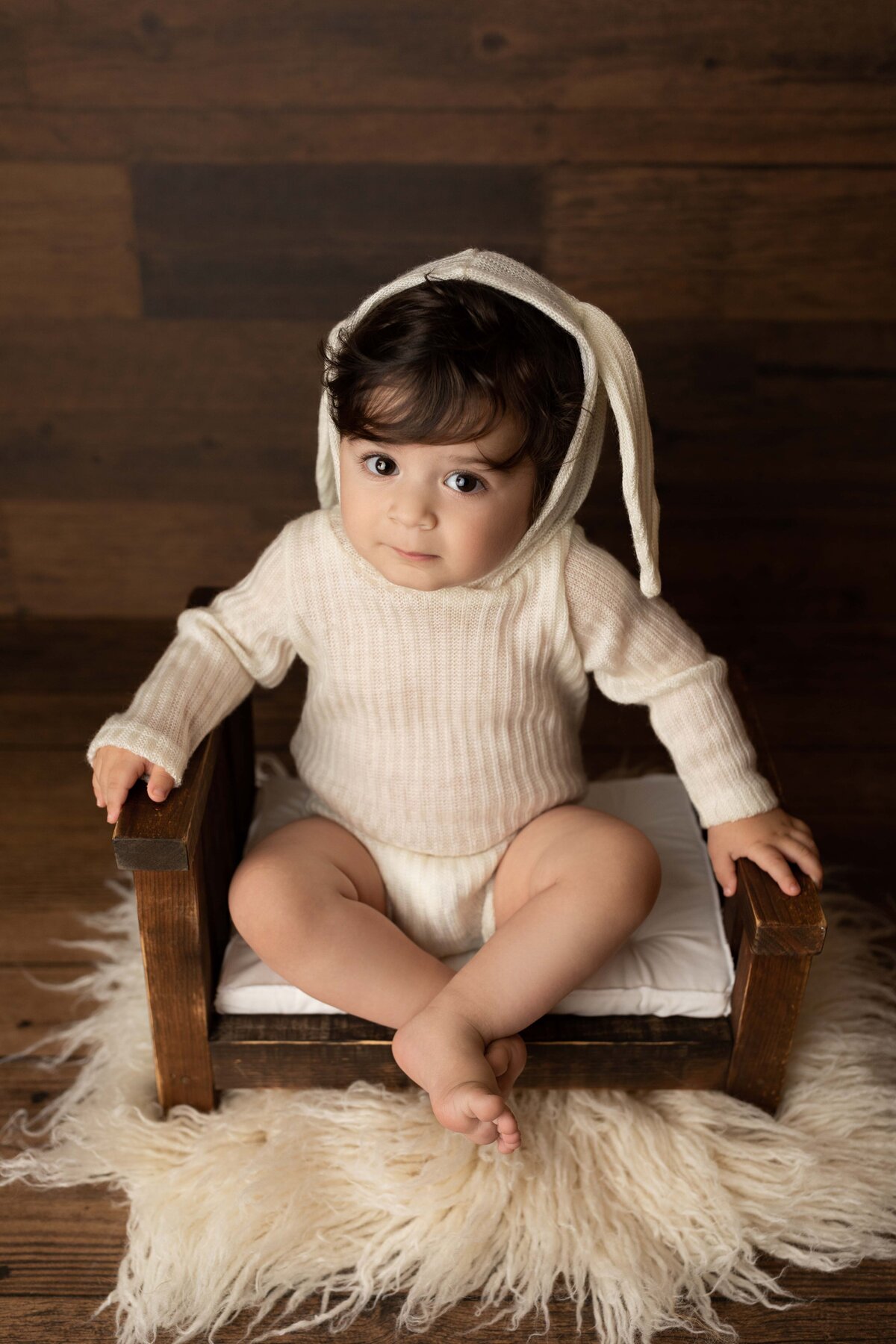 One-year-old sitting on little stool wearing a cream knit onesie with bunny ear hood. Baby is looking intently at the camera.