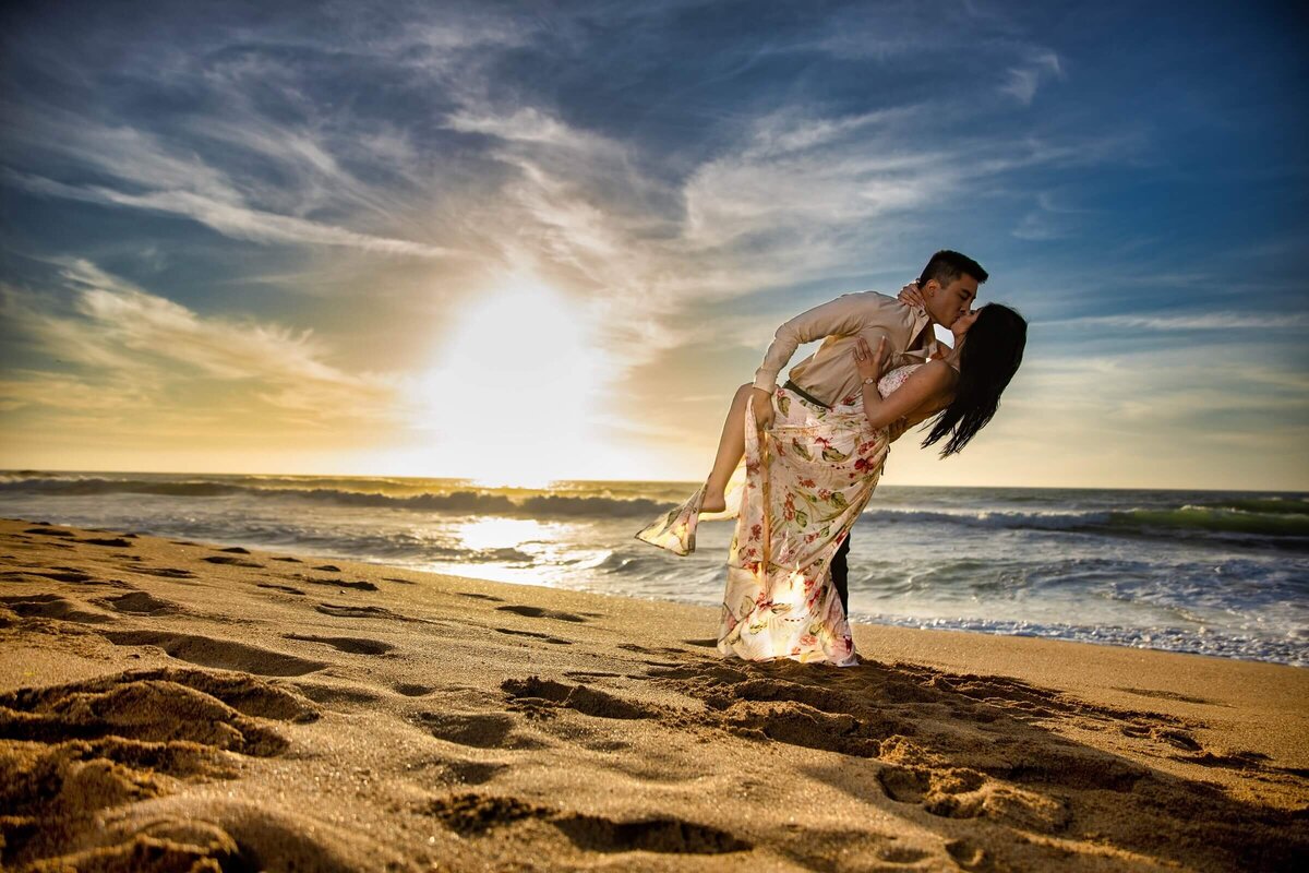 Engagement photo session of a couple on a beech with the man dipping the woman in front of the ocean and sunset.