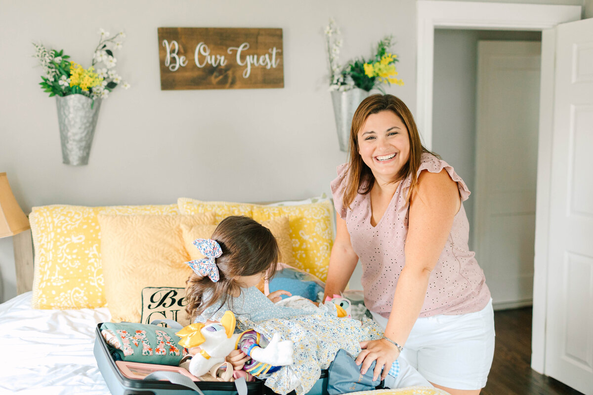 A smiling woman in a ruffled pink top and white shorts leans over a suitcase on a bed, while a young girl with a floral bow in her hair packs stuffed toys and clothes. The room features yellow pillows, wall-mounted flower arrangements, and a "Be Our Guest" wooden sign.
