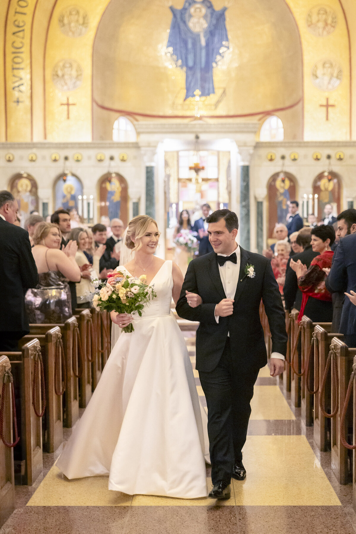 A bride in a white gown and a groom in a black tuxedo are joyfully walking down the aisle of a grand church, holding hands. They are surrounded by seated guests and ornate gold and blue decorations.