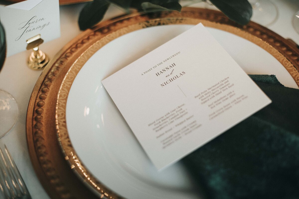 White squared place cards with black font set atop a dark green velvet napkin, white plate and bronze charger.