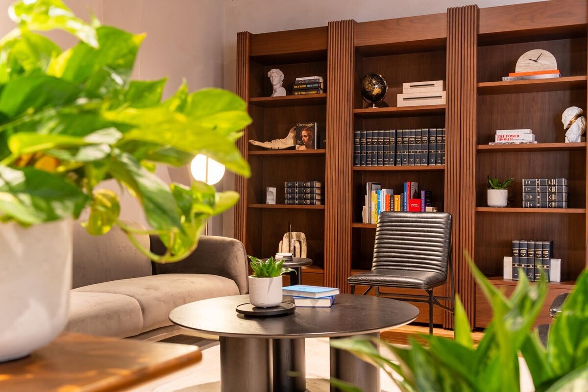 Lounge area with a wood bookshelf filled with books and decor, a gray sofa, a black accent chair, and greenery in the foreground.