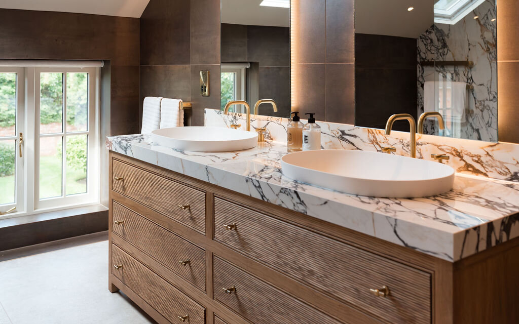 A modern bathroom with a sloped ceiling, featuring a double sink vanity with marble countertops and wooden cabinets. Two arched mirrors are mounted above the sinks, and large windows provide natural light. The room is finished with dark tiled walls and light tiled floors.