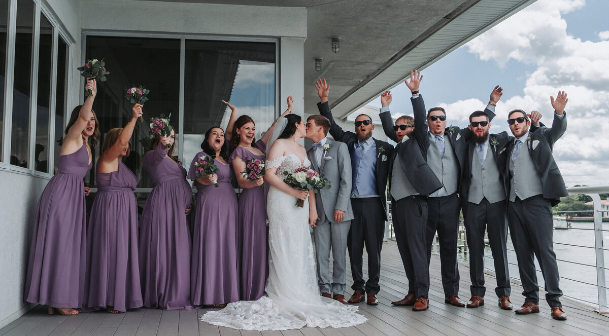 An excited wedding party, featuring bridesmaids in purple gowns and groomsmen in gray suits, celebrates with raised hands on a waterfront balcony at the Lesner Inn in Virginia Beach, Virginia, captured by Quinn Photography LLC.