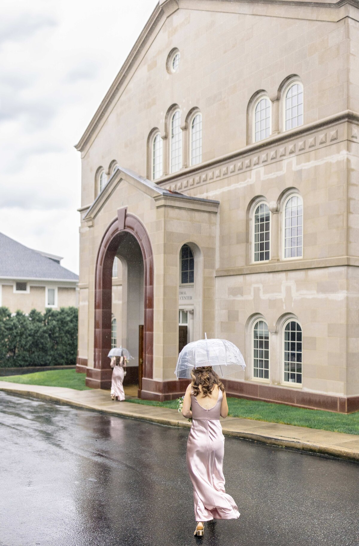 A woman in a pink dress walks on a wet road holding a transparent umbrella. She approaches a large beige building with arched windows and a red-brown entrance. Another person in pink stands under the entrance. Its cloudy and overcast.