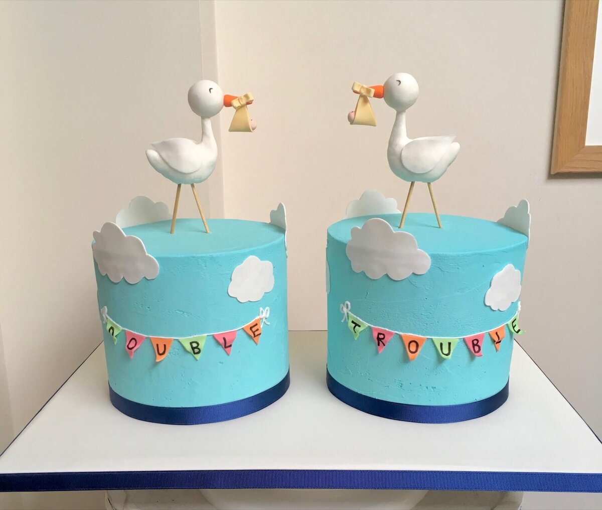 Two blue cakes that are almost identical with storks on top of each cake holding one baby each and buntin on the cakes, one saying "double" and the other saying "trouble"