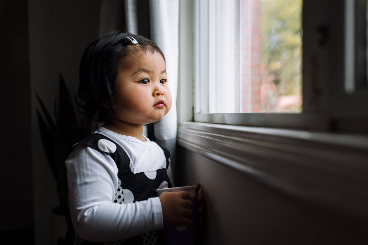 toddler girl standing by window at home holding snack and looking outside with serious expression