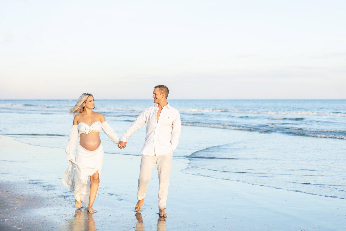 Expecting parents holding hands as they walk along the beach