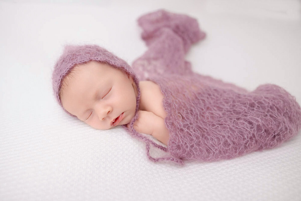 two-week baby girl posed lying on white blanket and wearing a purple bonnet and covered in a lacy purple baby wrap.
