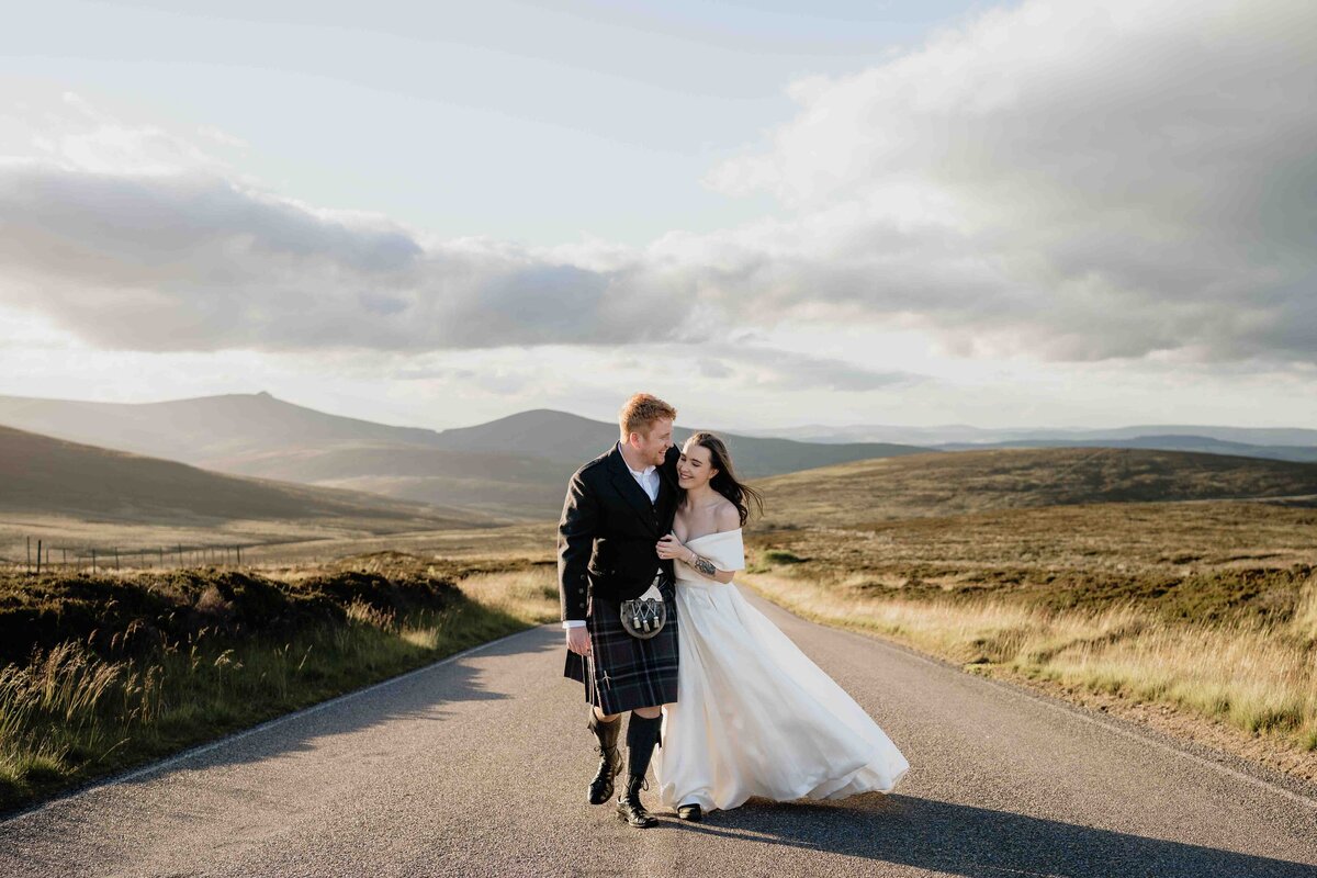 A couple walks hand in hand on a scenic road near Aberdeen, surrounded by the lush, rolling hills of Cairn o’ Mount. Captured by their wedding photographer, the man wears a kilt, and the woman a flowing white dress. The partially cloudy sky casts soft light on the picturesque landscape.