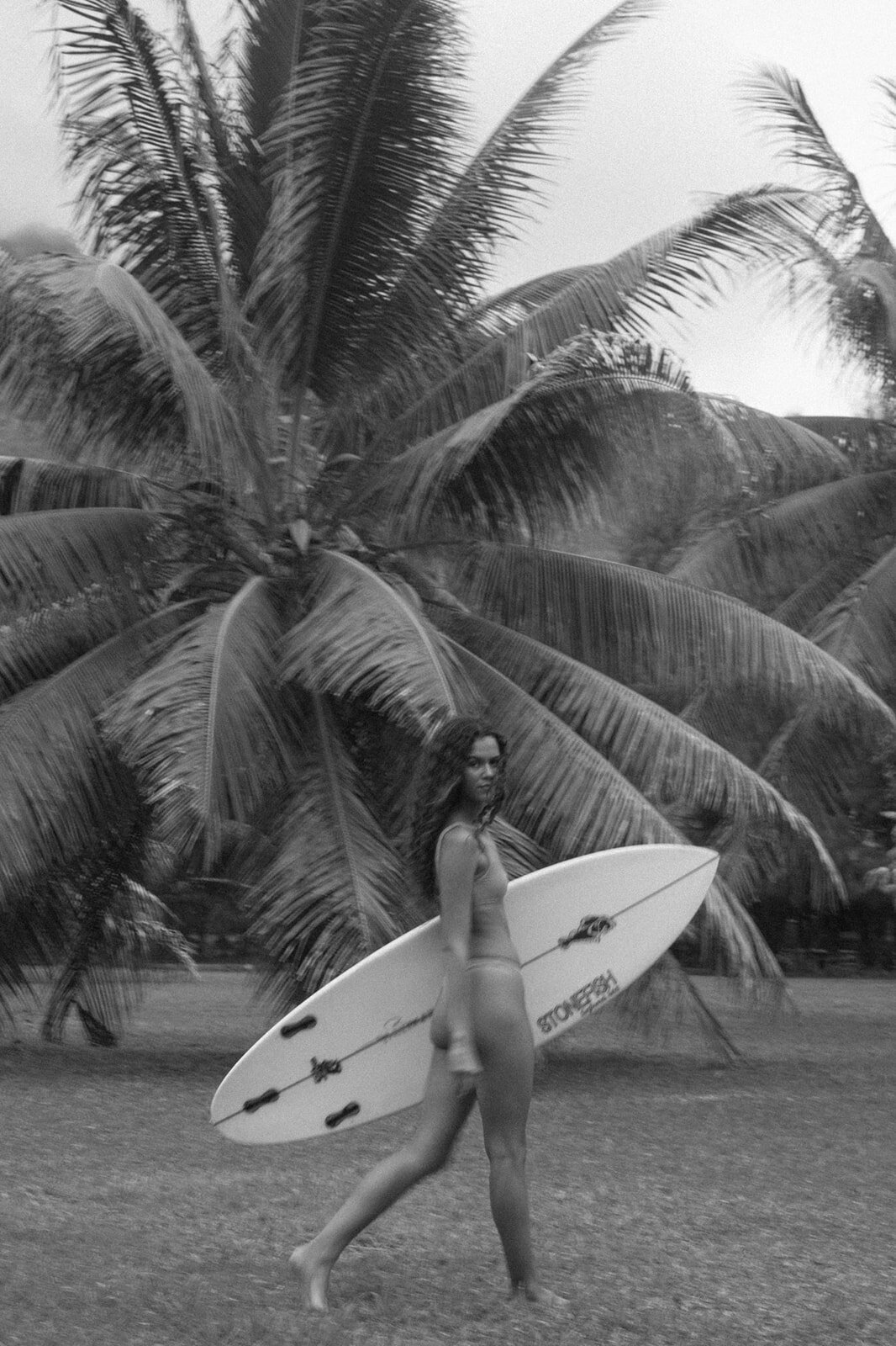 Black and white image of a female surfer holding a surfboard, walking on grassy ground with tall tropical palm trees in the background, evoking a beach and surfing lifestyle.

