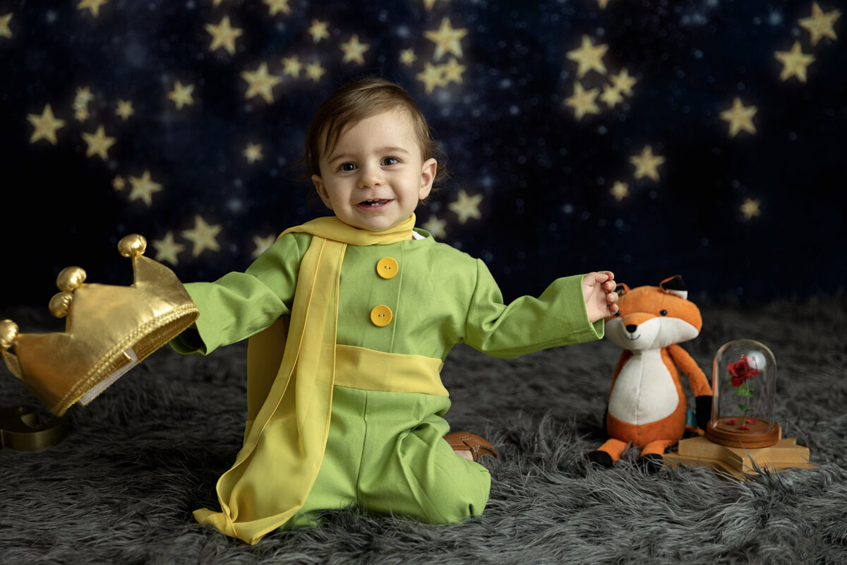 A happy toddler boy in a green prince costume and crown happily sits on a grey blanket with a stuffed fox for a New Jersey Milestone Photographer