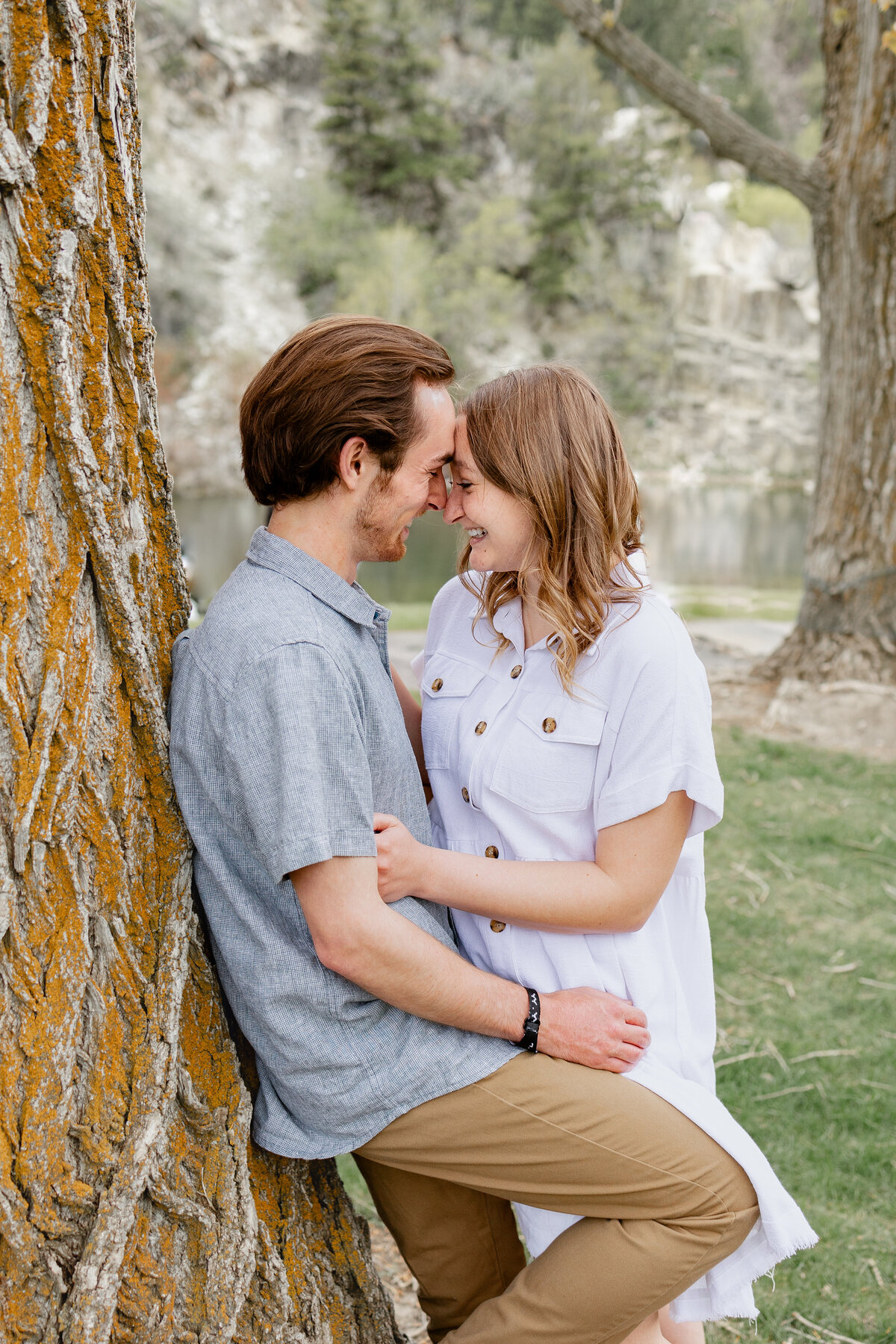 A couple stands together, lovingly leaning against a tree with textured bark, with a mountain and river scene in the background of Vivian Park, Provo Canyon