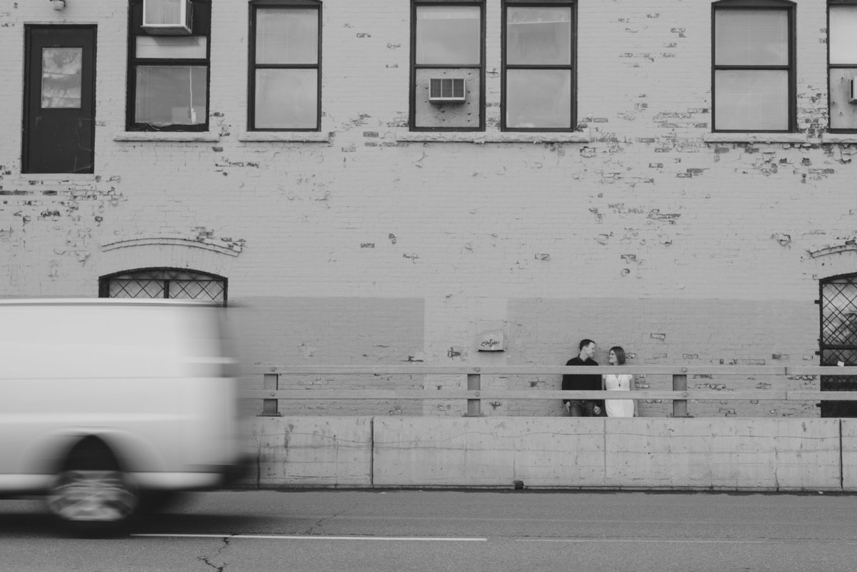 Couple standing against old factory wall with white truck driving past