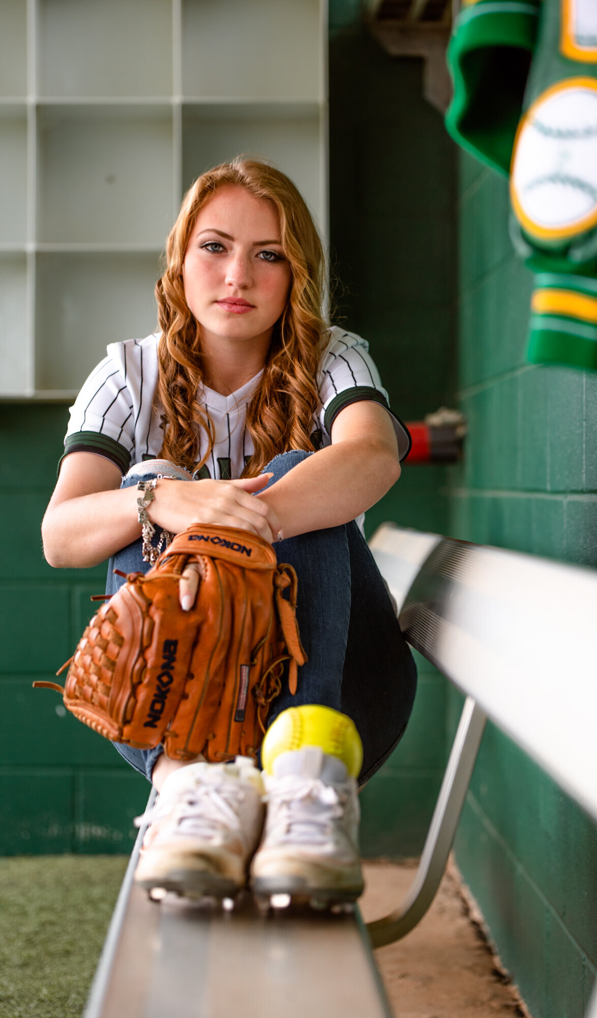A softball player with red hair sits on a dug out bench with her cleats, ball, glove, and letterman.