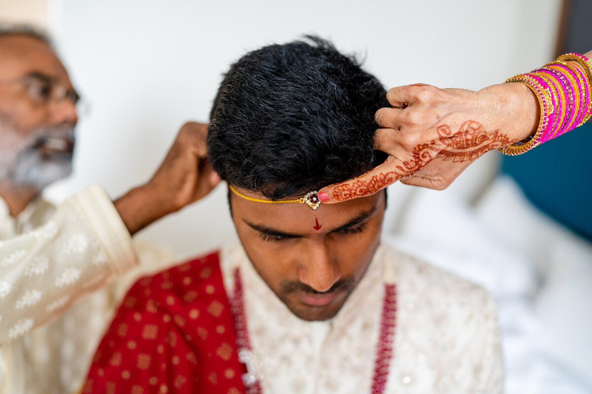 A groom dressed in traditional Indian wedding attire receives a ritual adornment on his forehead, applied by a person with henna-decorated hands.