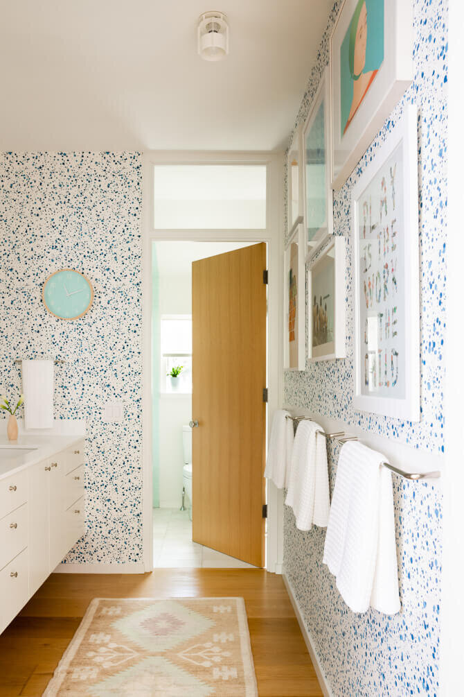 Interior designer in Hingham creates a modern and playful bath with a white vanity, Hinson splatter wallpaper, colorful art, a natural oak door, and blue accents.