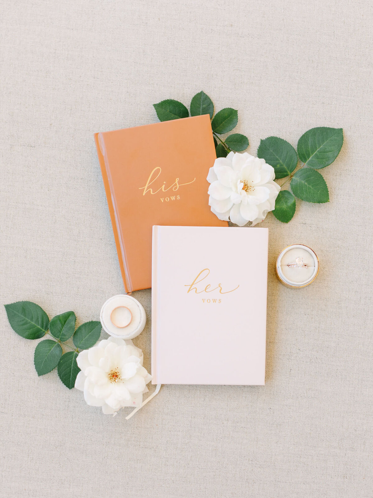 Two vow books, labeled "his" and "her," are surrounded by white roses, green leaves, and a ring box on a beige background.