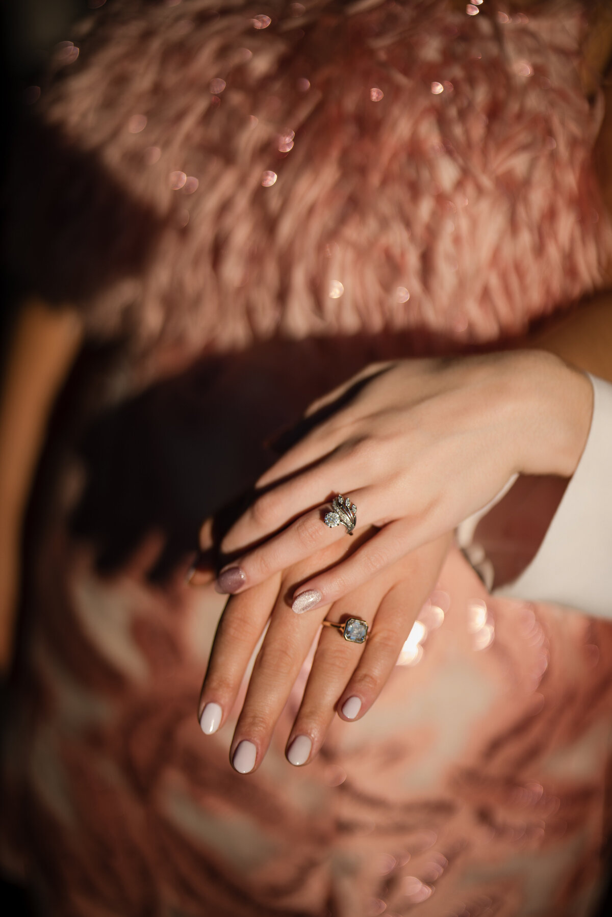 Close-up of two hands with manicured nails, wearing two sparkling rings, resting on a sequined, textured pink fabric.