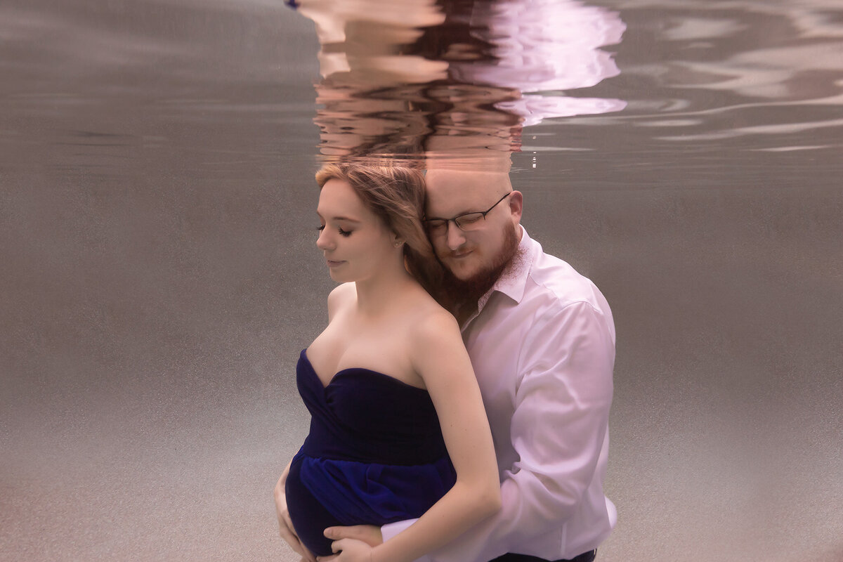 A serene underwater maternity photo captured by a Dallas underwater pregnancy photographer. A couple shares a tender moment beneath the surface, the man holding his partner close from behind with a peaceful, loving expression. The woman wears a flowing deep blue gown, her hands gently cradling her belly, enhancing the ethereal mood of the scene. The soft, neutral background complements the couple's connection, emphasizing the tranquility and beauty of this underwater maternity photoshoot.