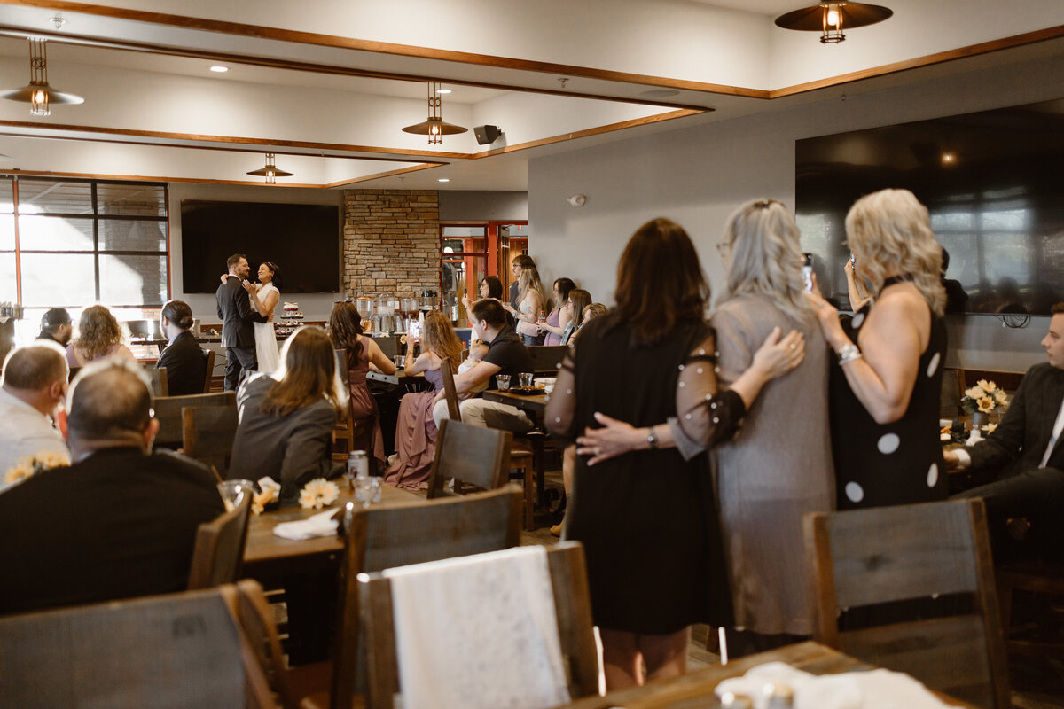 A bride and groom share their first dance in a cozy indoor reception venue while seated guests and standing family members watch and record the moment.