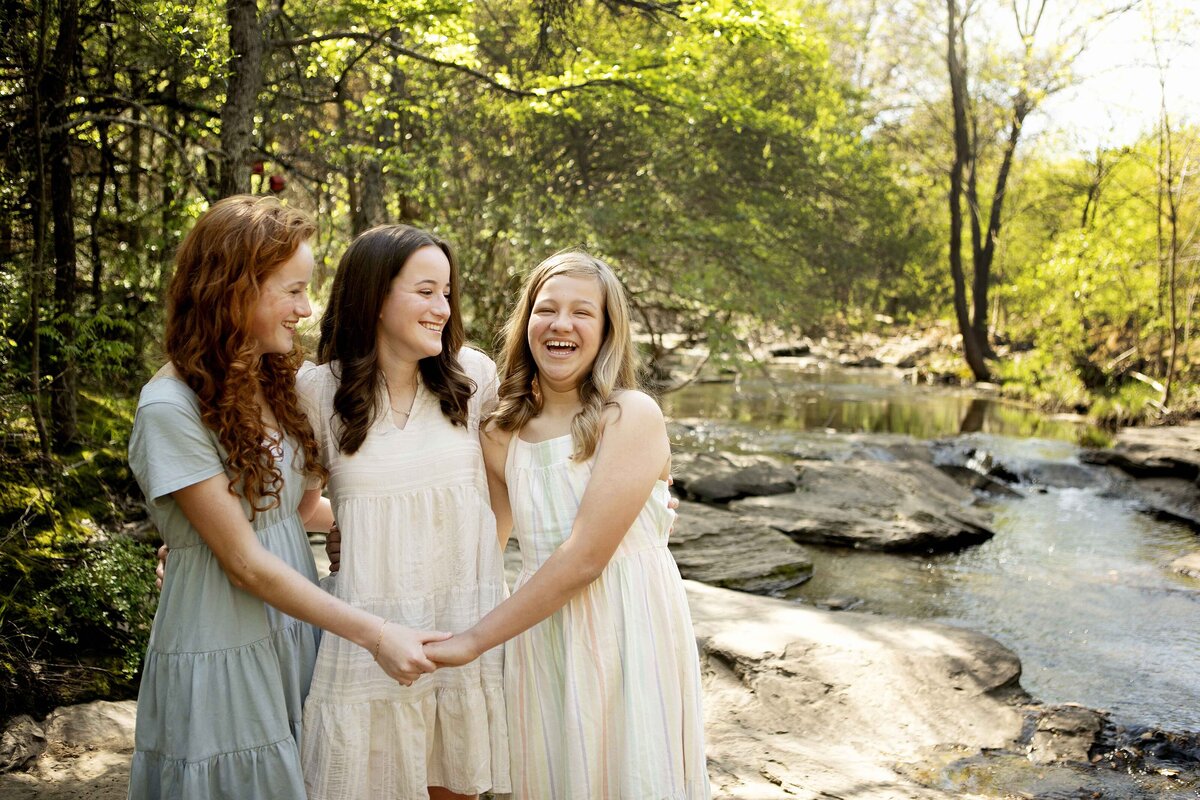 The three sisters giggling and playfully interacting by the creek in Flower Mound, capturing a fun moment in their photoshoot