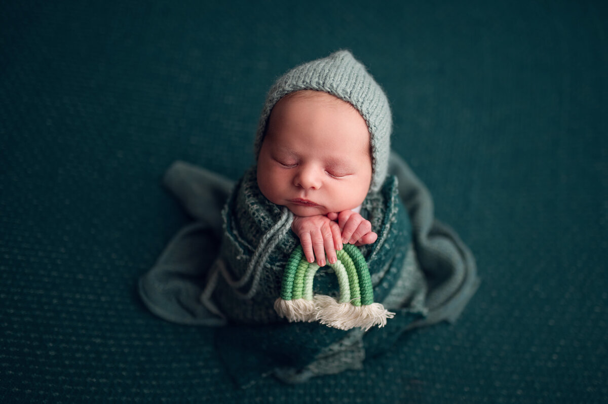 Green themed infant portrait showing sleeping baby on a green background. He is wrapped in a green, knit blanket and coordinating hat while holding a tiny green rainbow.