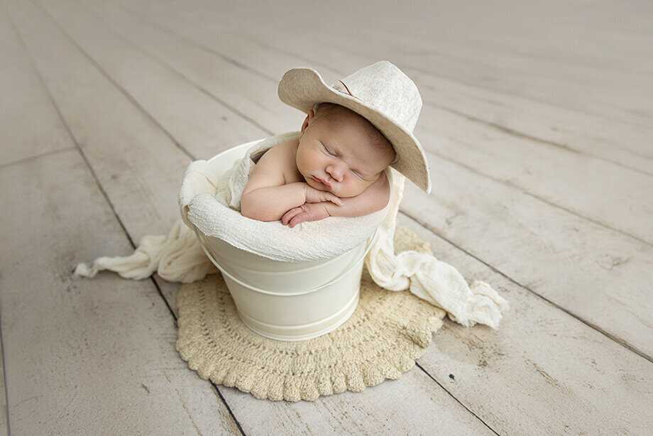 newborn boy posed in a cream bucket with a matching cream colored cowboy hat.