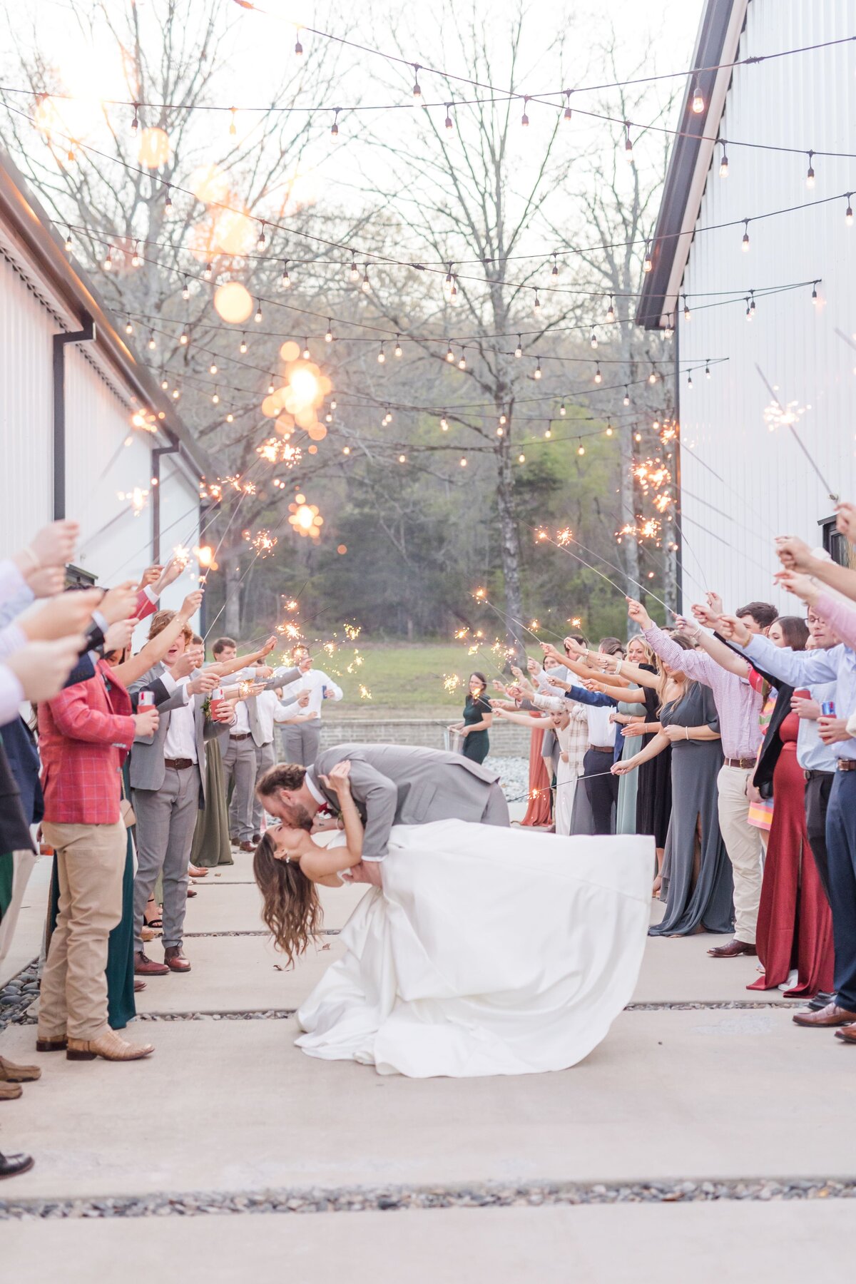 A groom in a grey suit dips his bride for a kiss under sparklers and market lights