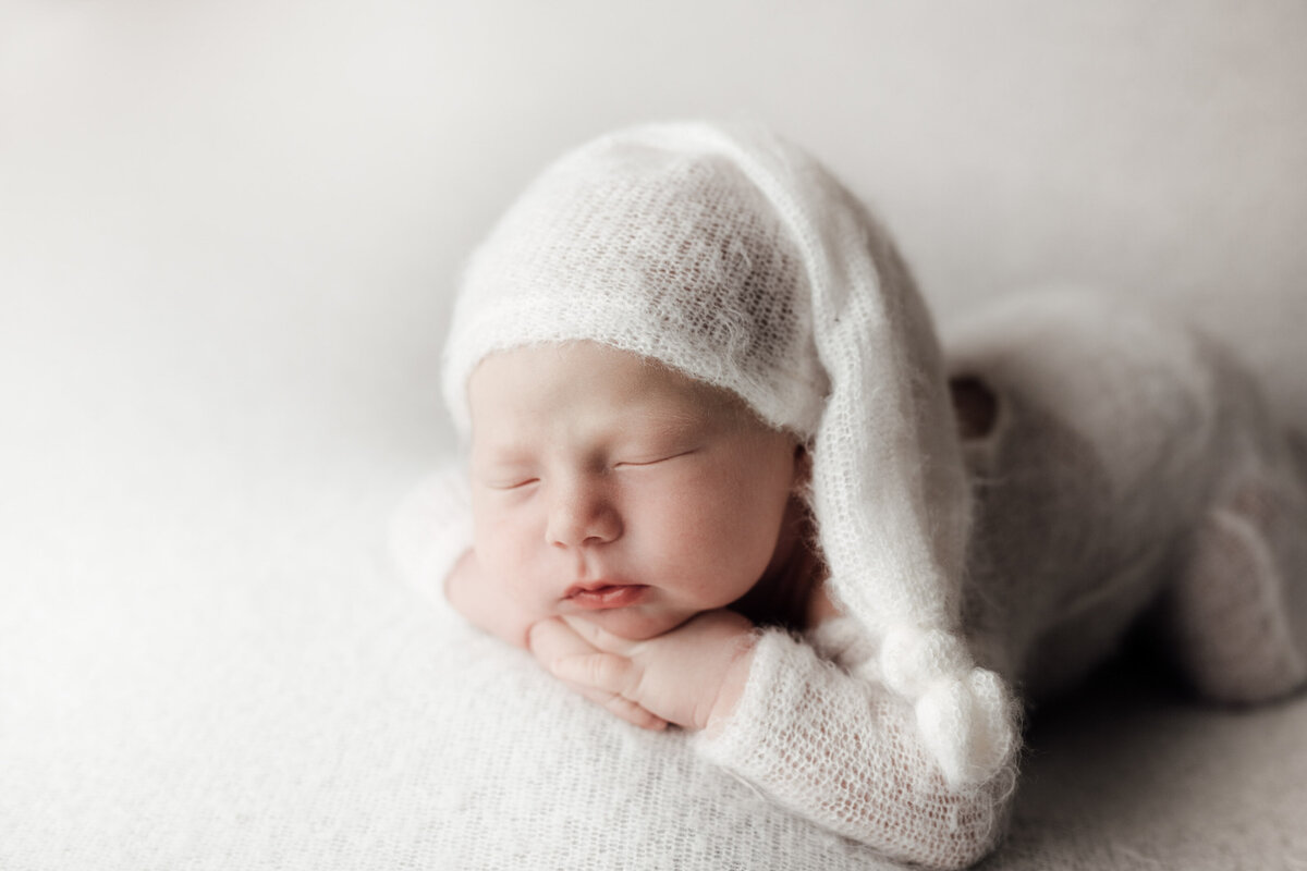 Baby boy in cream kit onesie with his hands folded under his chin. baby is wearing a matching long knit cap draped to his elbow.