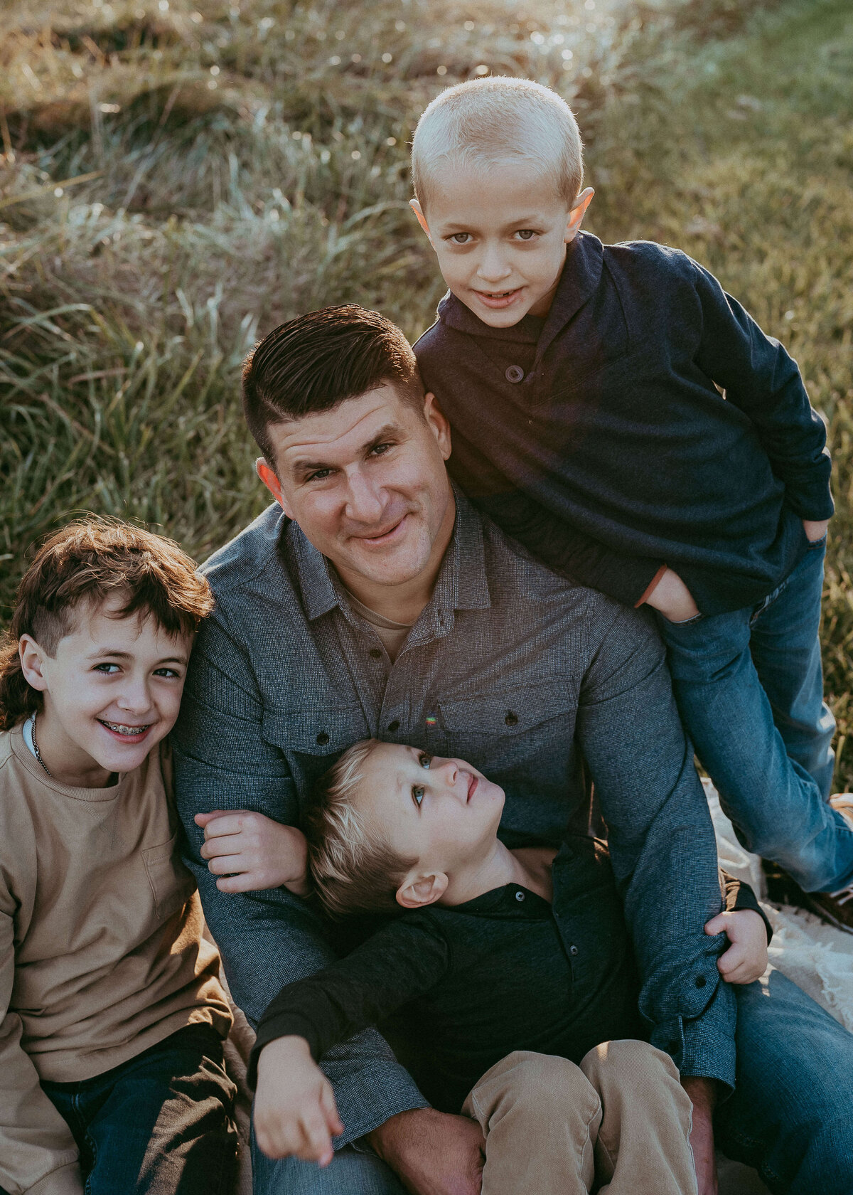 Three boys adoringly hug on their large football player dad's body as they all sit on a blanket in tall grass on their family farm wearing navy blue and denim colors.