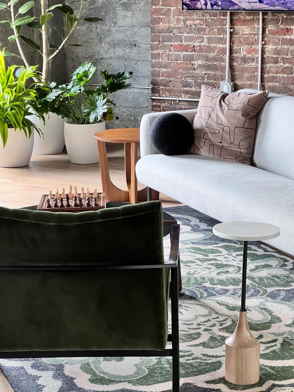 Styled seating vignette with a white sofa, patterned pillows, wood side tables, indoor plants, and a brick accent wall.