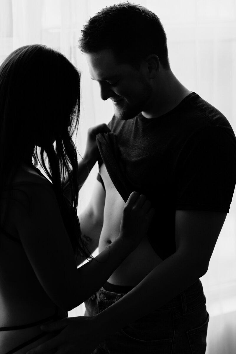 a black and white image of a wife slowly removing the man's shirt. the man is smiling.