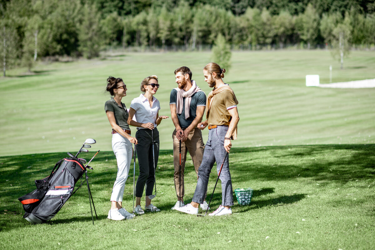 Four people stand on a golf course, talking and holding golf clubs. A golf bag is nearby on the grass. They're dressed in casual golf attire, and a basket of golf balls is visible. Trees and the fairway are in the background.