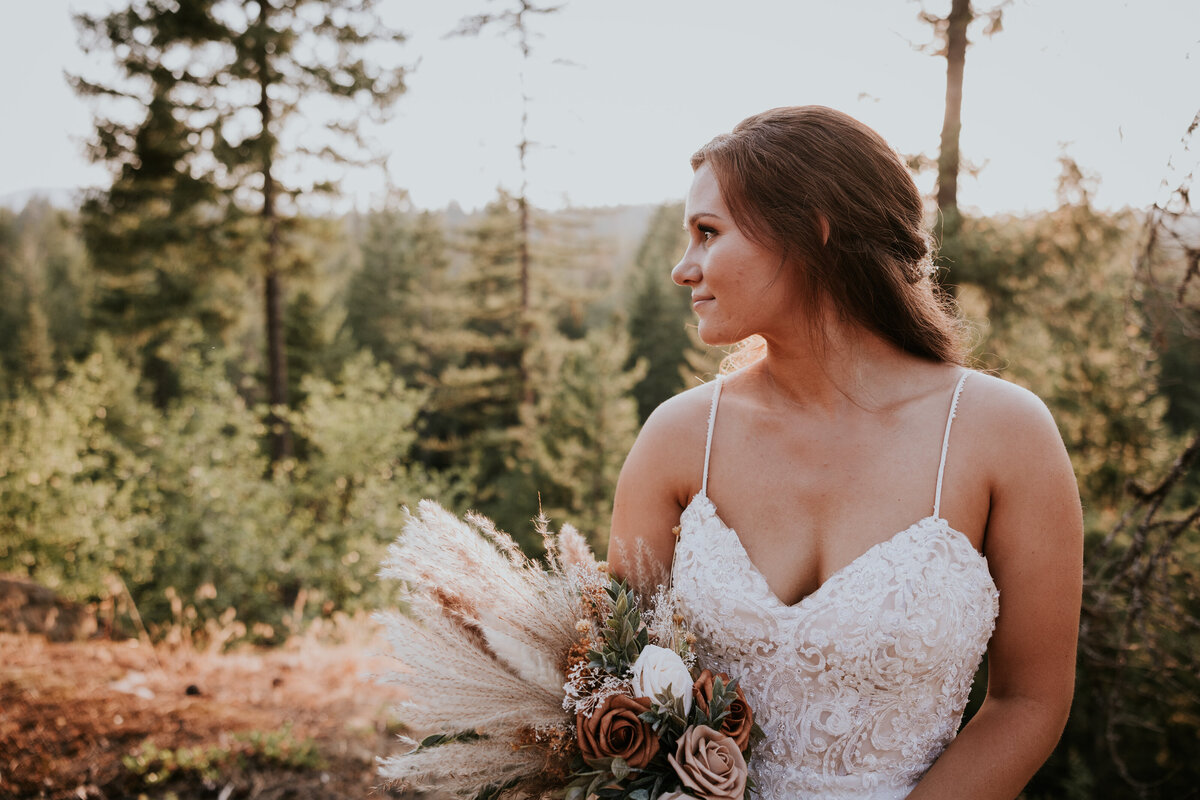 Bride looking out at forest during sunset holding bouquet.