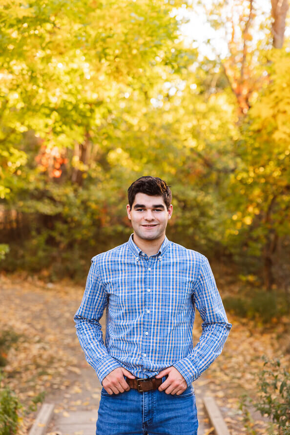 Boy smiles at camera with hands in pockets with fall foliage behind him.