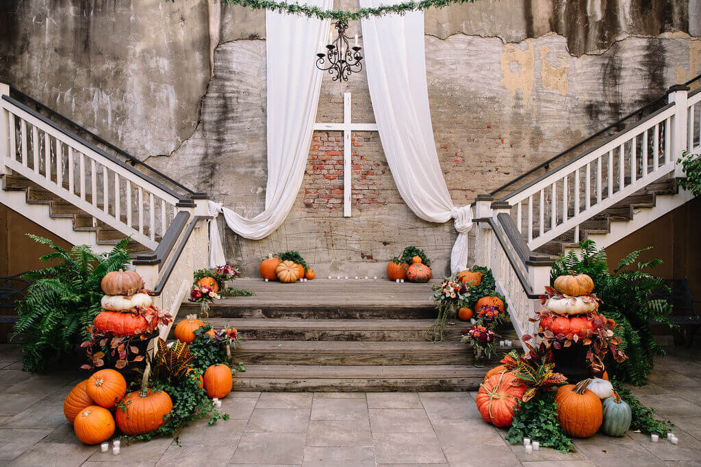 Several vibrant floral bouquets featuring roses, eucalyptus, and other colorful flowers are arranged on wooden steps with pumpkins and vines. A white cross hangs on a brick wall at the back.