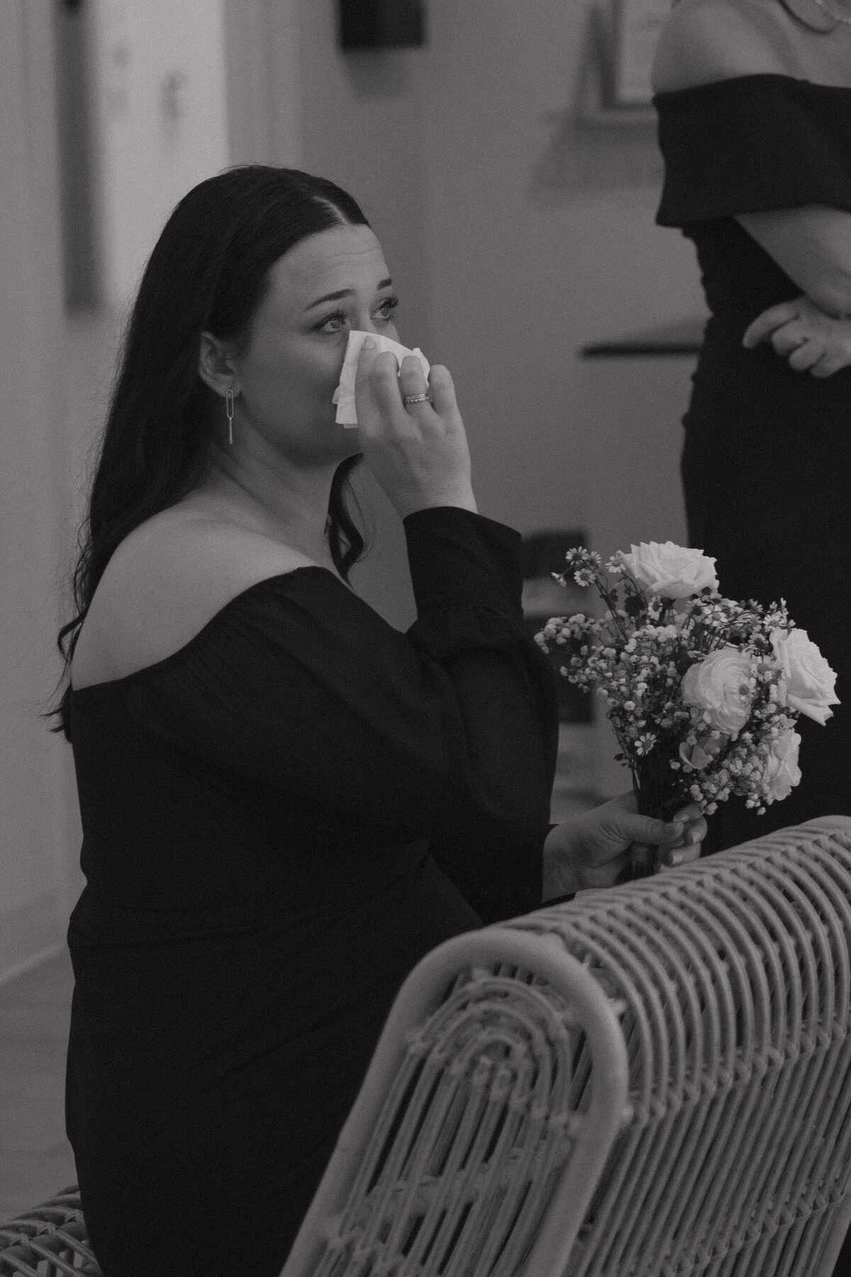 A bridesmaid in a long sleeve off the shoulder black dress sits backwards in a wooden chair wiping a tear from her eye with a tissue in her right hand. In her left hand she holds a small bouquet of flowers with baby's breath, roses and peonies. 