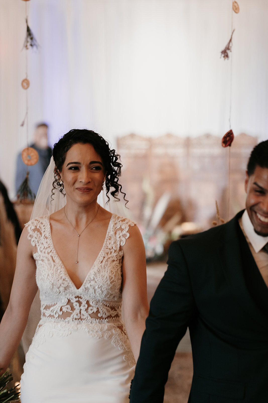 Bride walking down the aisle after the ceremony, smiling with emotion while holding her husband's hand, surrounded by a softly lit background.