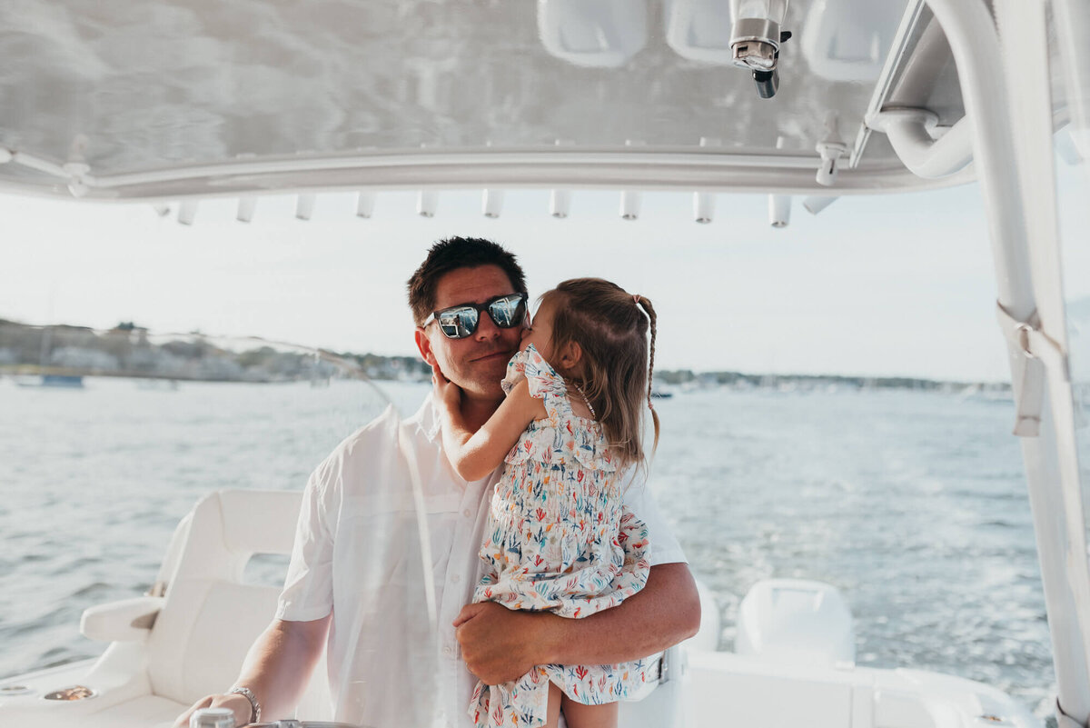 A father holds his young daughter as he drives their boat, she kisses him on his cheek during their photoshoot with Allison Wolf, Boston Family Photographer
